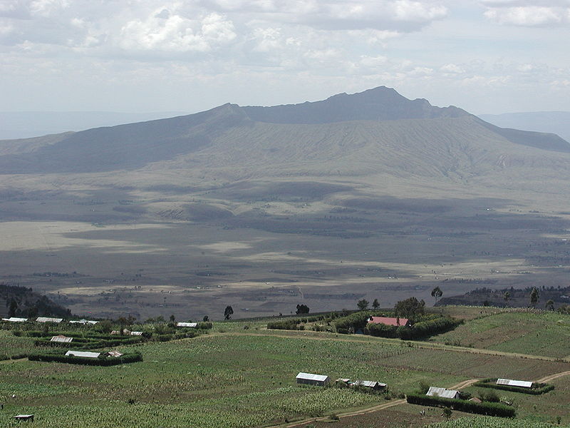 A wide, low volcano, seen from nearby highlands, with weathered sides covered in scrub brush and a large caldera